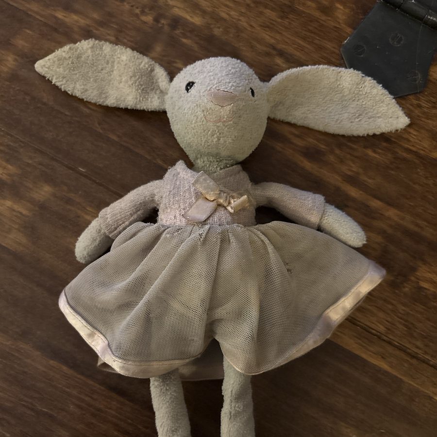 A well-loved grey toy bunny in a lilac cardigan and tulle skirt, photographed on a kitchen table